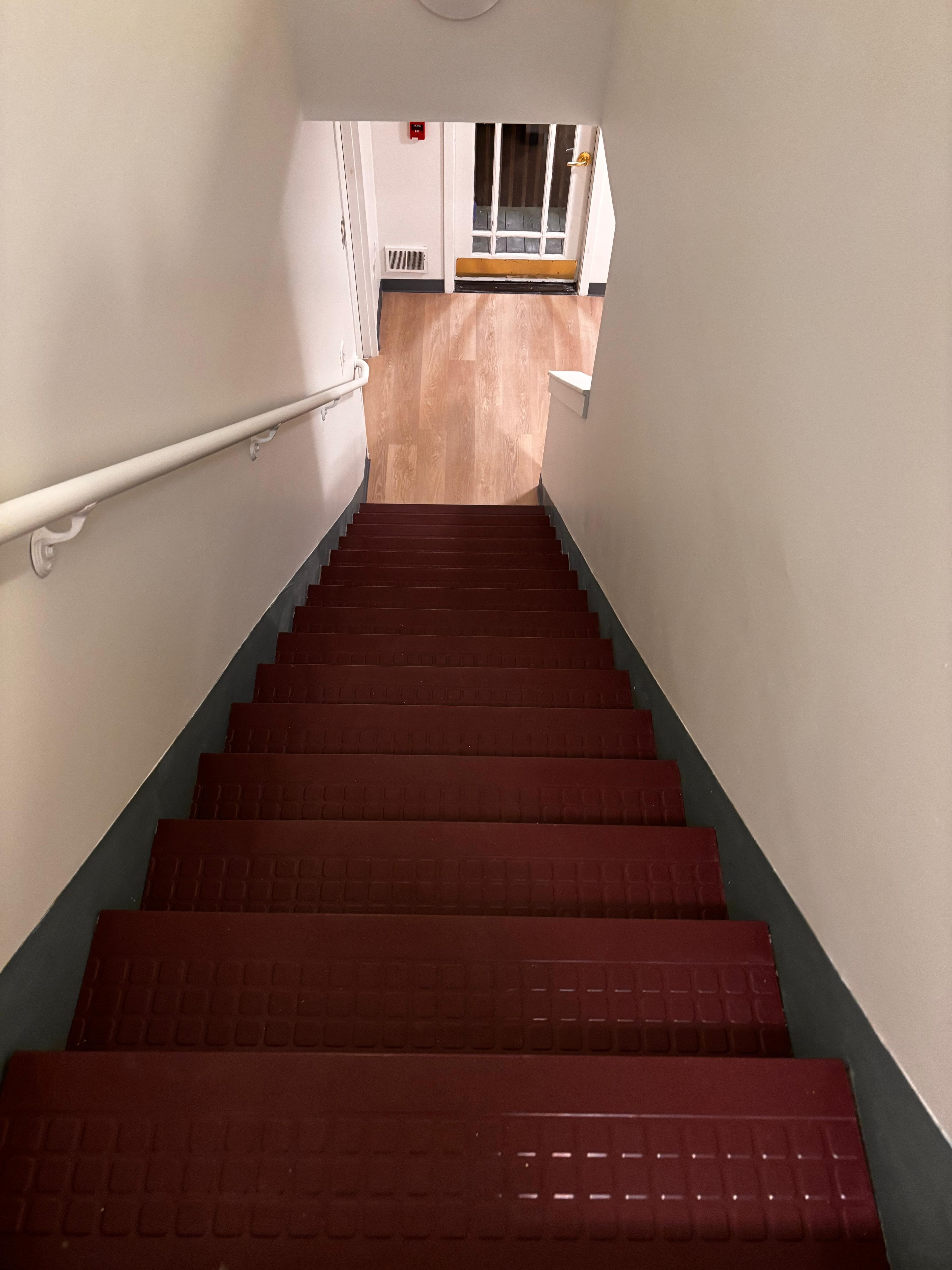 Looking down dark red stairs with a white handrail toward a light wood floor.