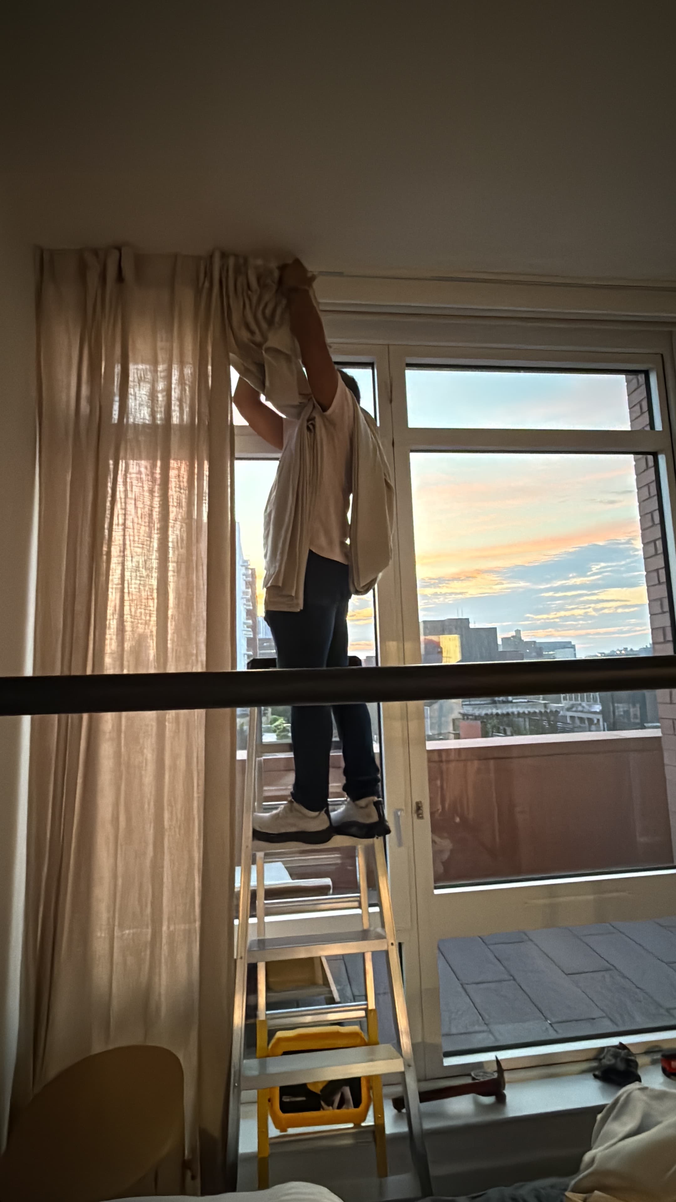 Person on a stepladder hanging beige curtains in front of a window at sunset.