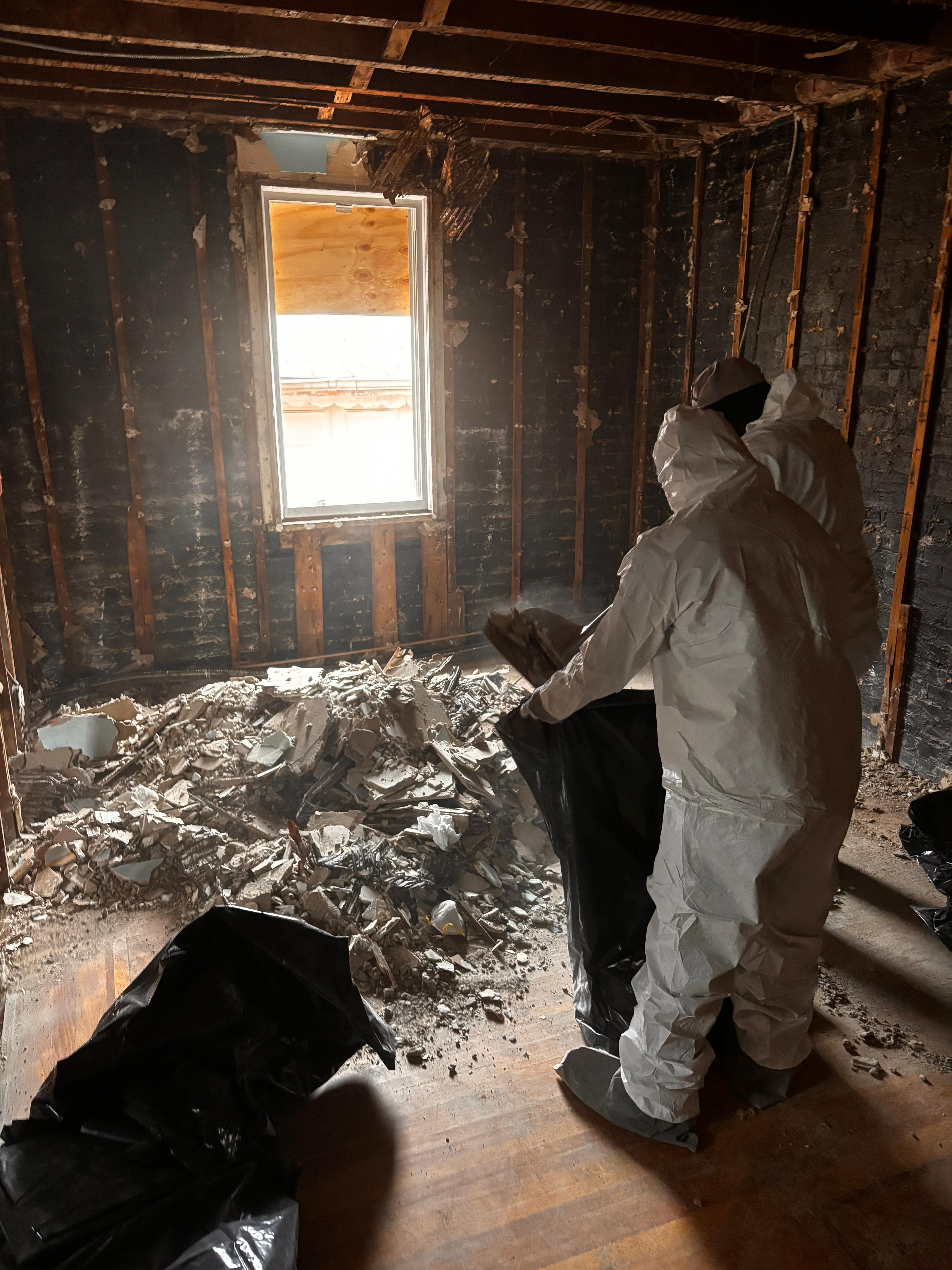 Workers in white hazmat suits clearing a pile of debris from a room under renovation.