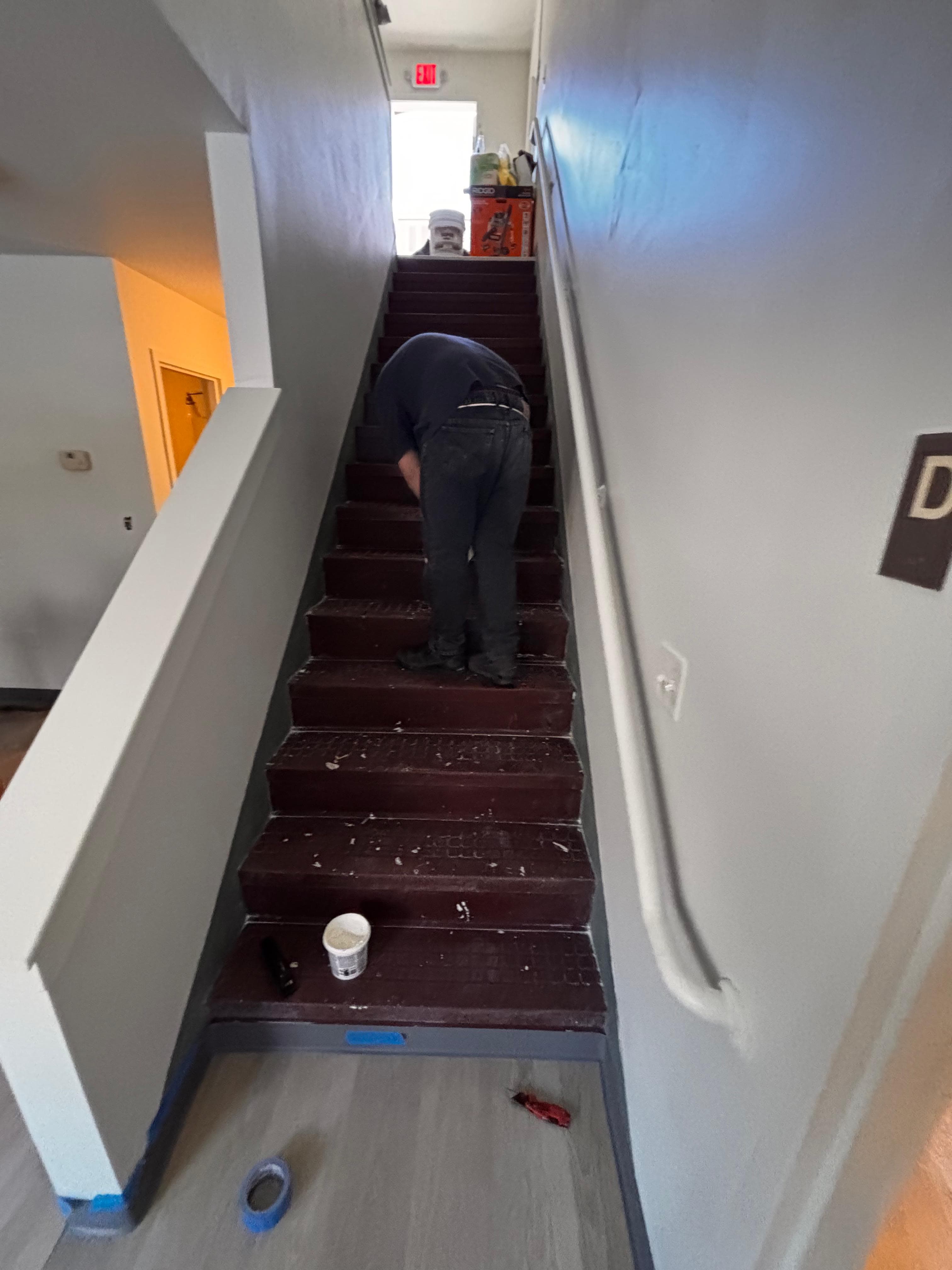 A person bent over working on dark brown stairs in a light gray indoor stairwell.