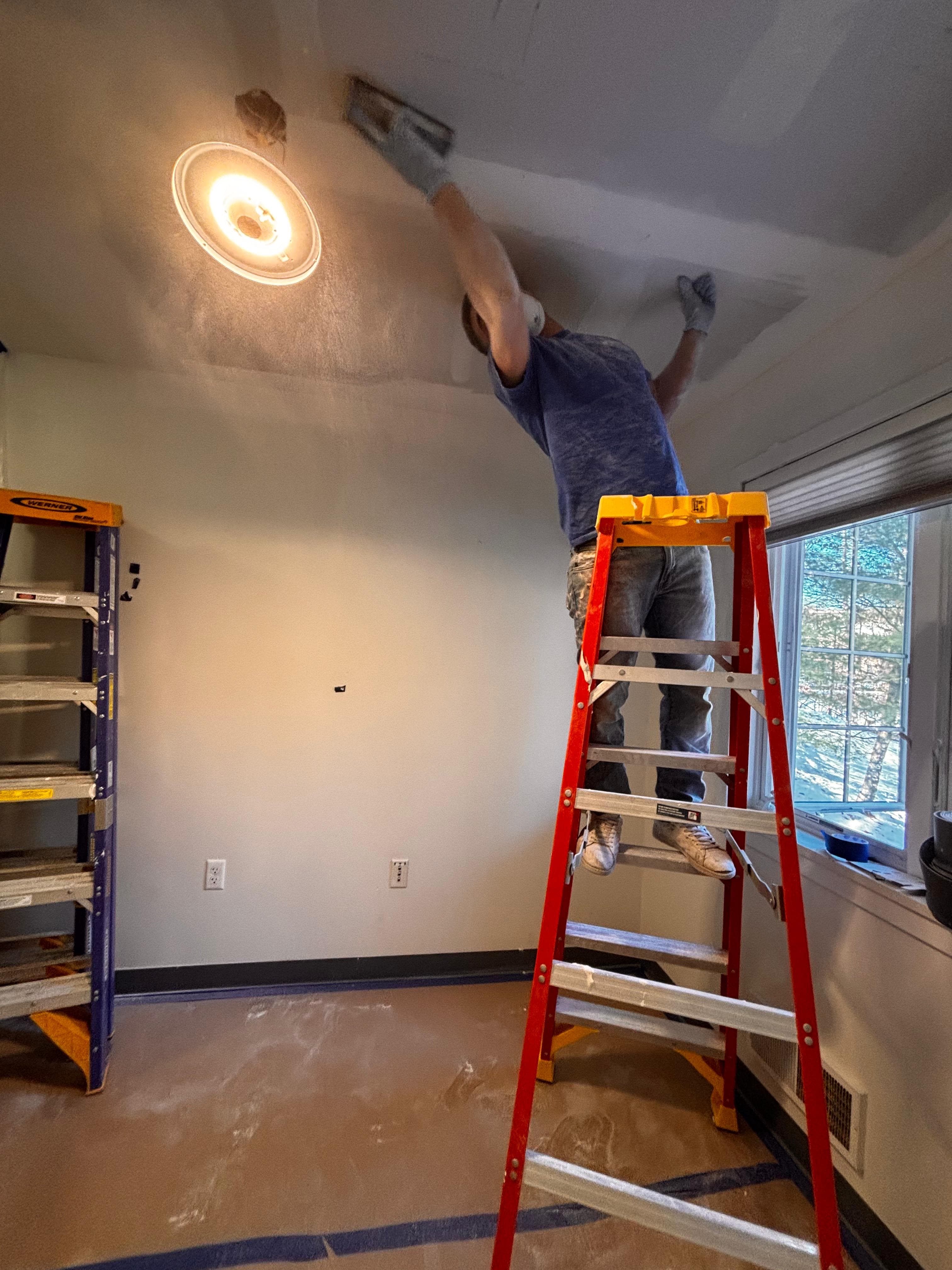 A worker on a red ladder repairs a ceiling in a room undergoing home renovation.