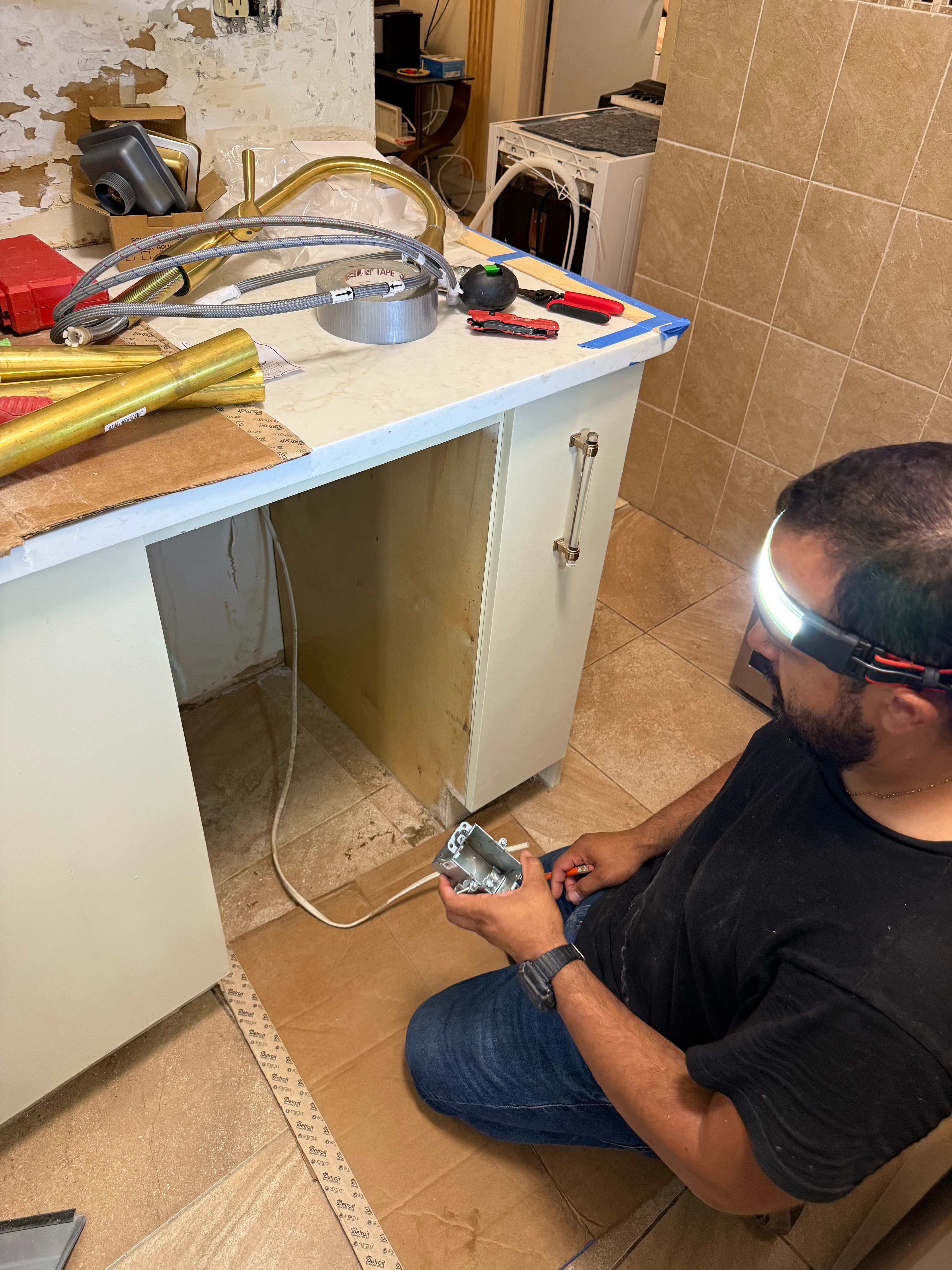 Man wearing a headlamp holds a metal electrical box while working on a kitchen renovation.