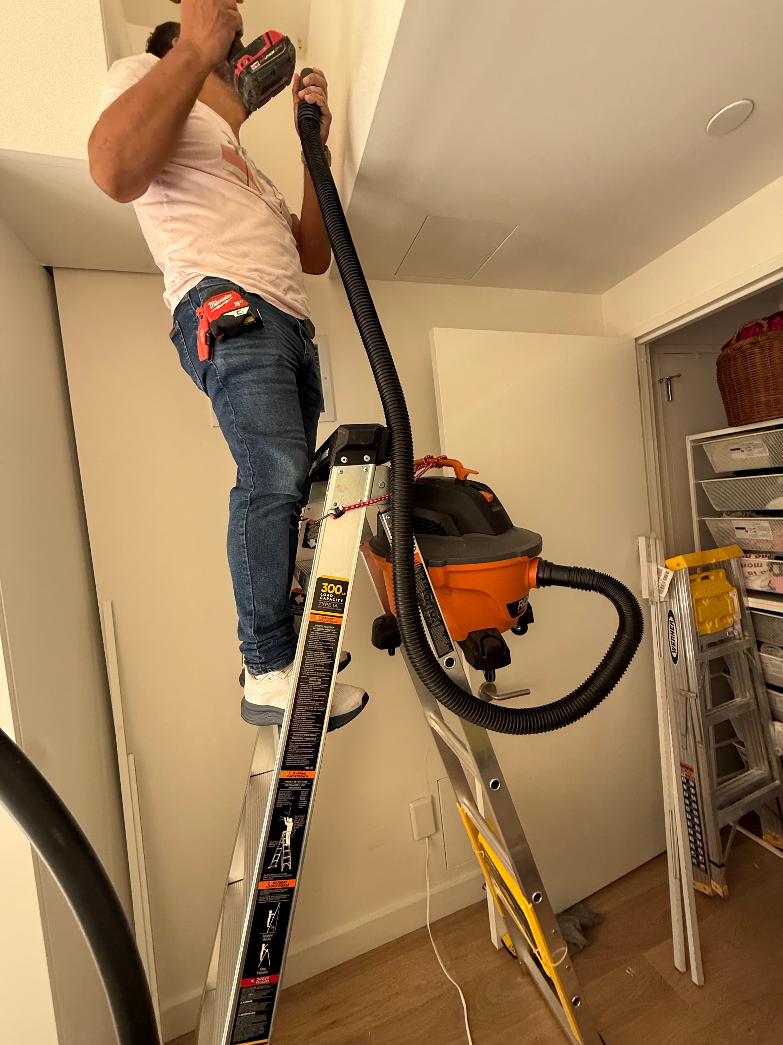 Man on ladder drills into ceiling while using a shop vac to catch dust.