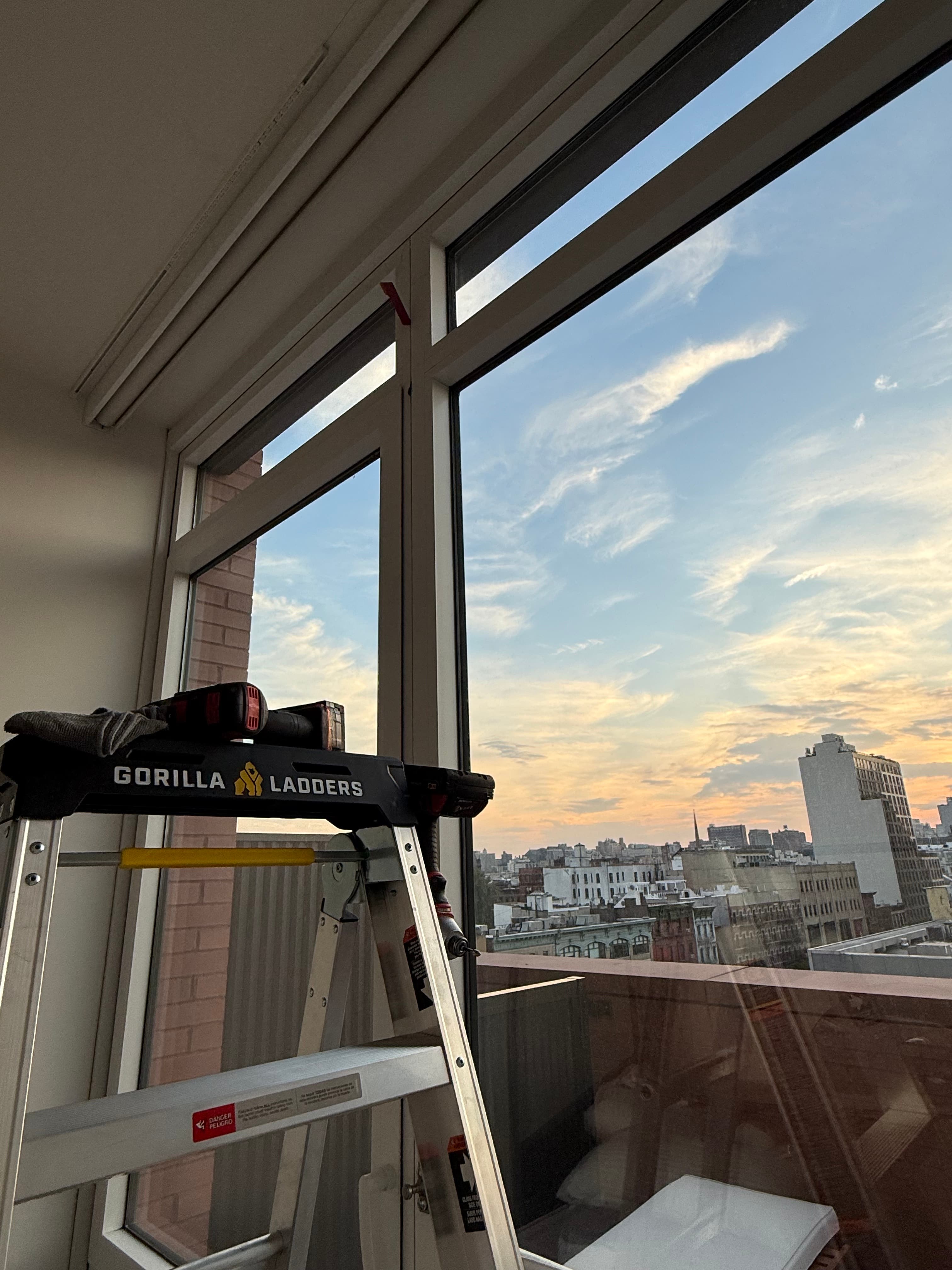 Gorilla ladder with a drill sits before a window overlooking a city skyline at sunset.