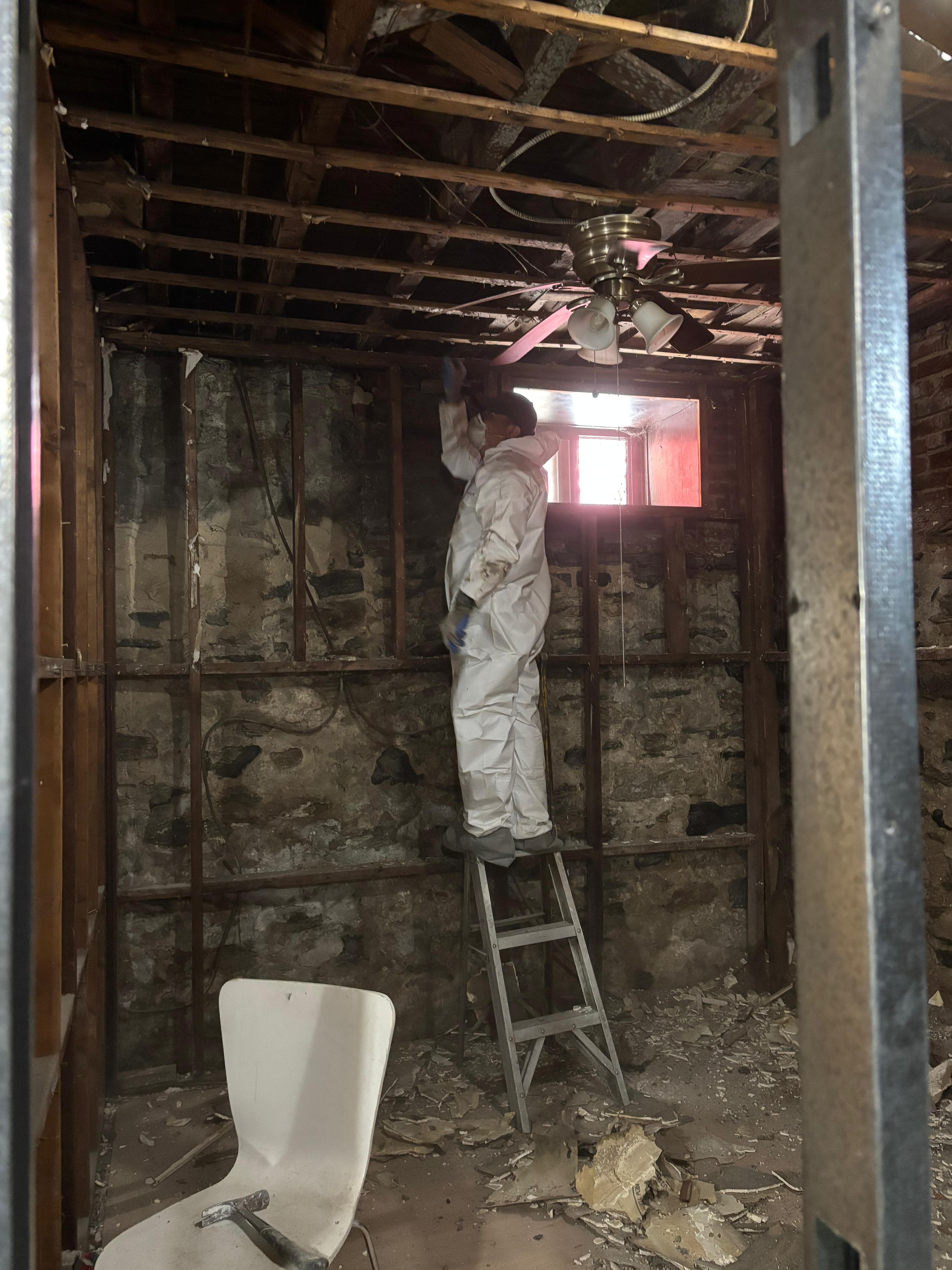 Worker in a white protective suit on a ladder renovating stone walls and exposed ceiling.